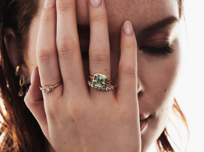 Close up of Model with Eyes Closed and Right Hand on Face Wearing 3 Rings Including 3.80 Carat Bezel Set Asscher Cut Green Tourmaline Ring and Cymbeline Ring with Round Diamonds from Vale Jewelry