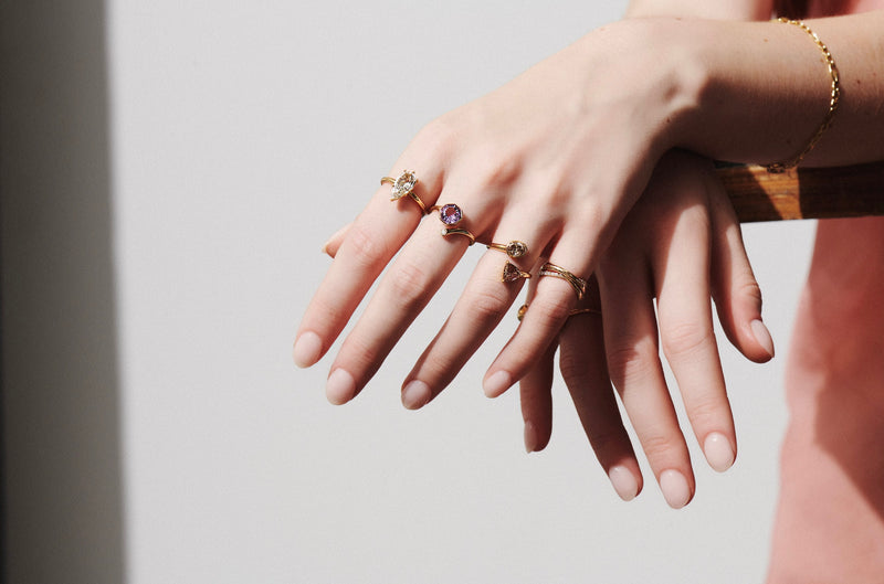 Close up of Model's Hand Hanging Over a Wooden Shelf Wearing One-of-a-Kind 1.50 Carat Step Cut Pear Diamond Solitaire Ring, Toi et Moi Champagne Diamond Ring, and Sintra Rolling Ring