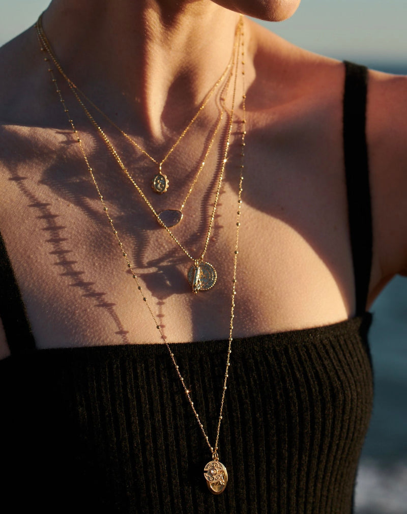 Model in Sunlight on Beach with Necklaces Layered on Back with Starlight Amulet Necklace, Protection Medallion Necklace and Figa Amulet Necklace from Vale Jewelry