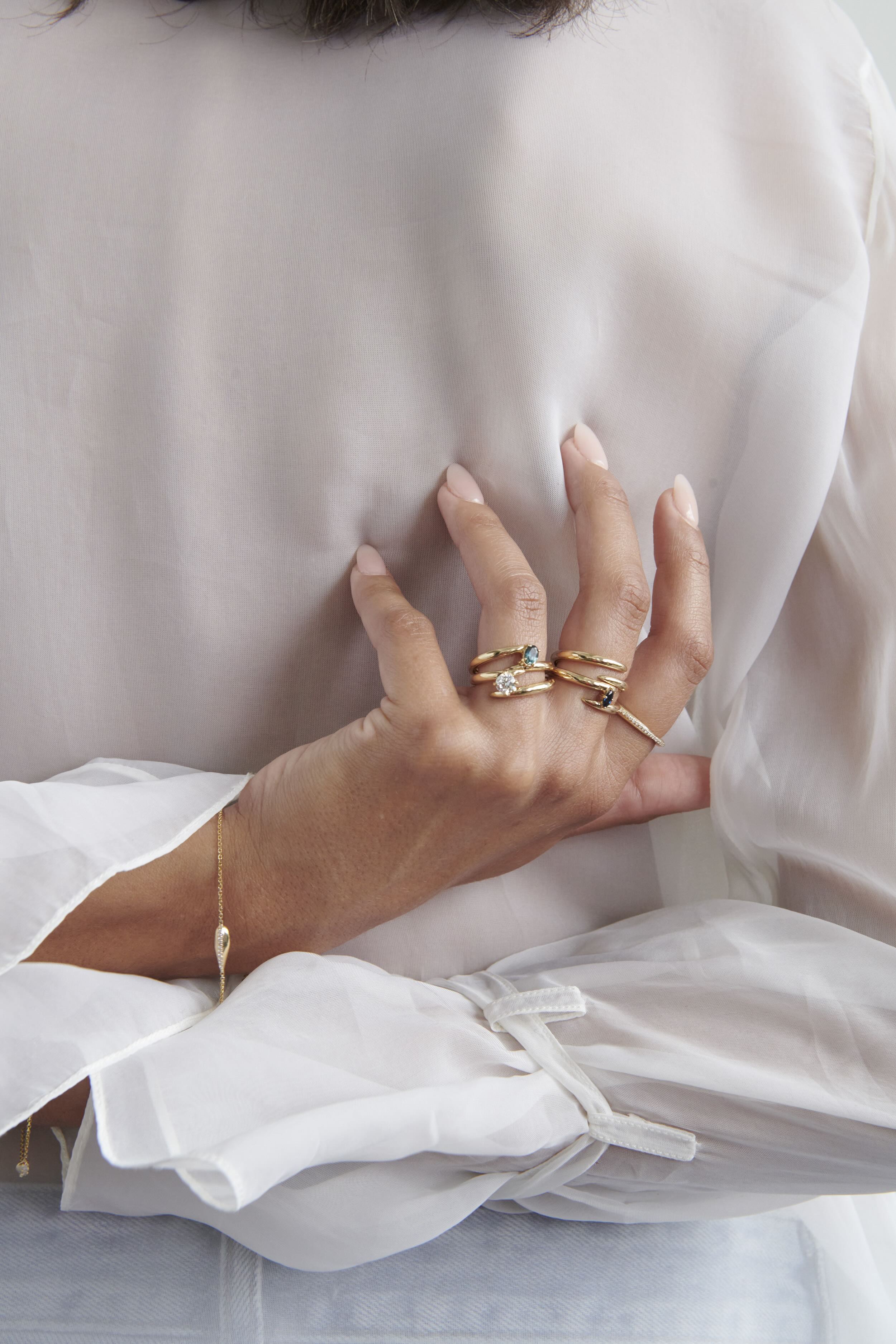 Close-up of Hand Model Wearing Vale Jewelry Swoop Marquise Cut Sapphire Bypass Ring with Multiple Gold Rings Against White Shirt