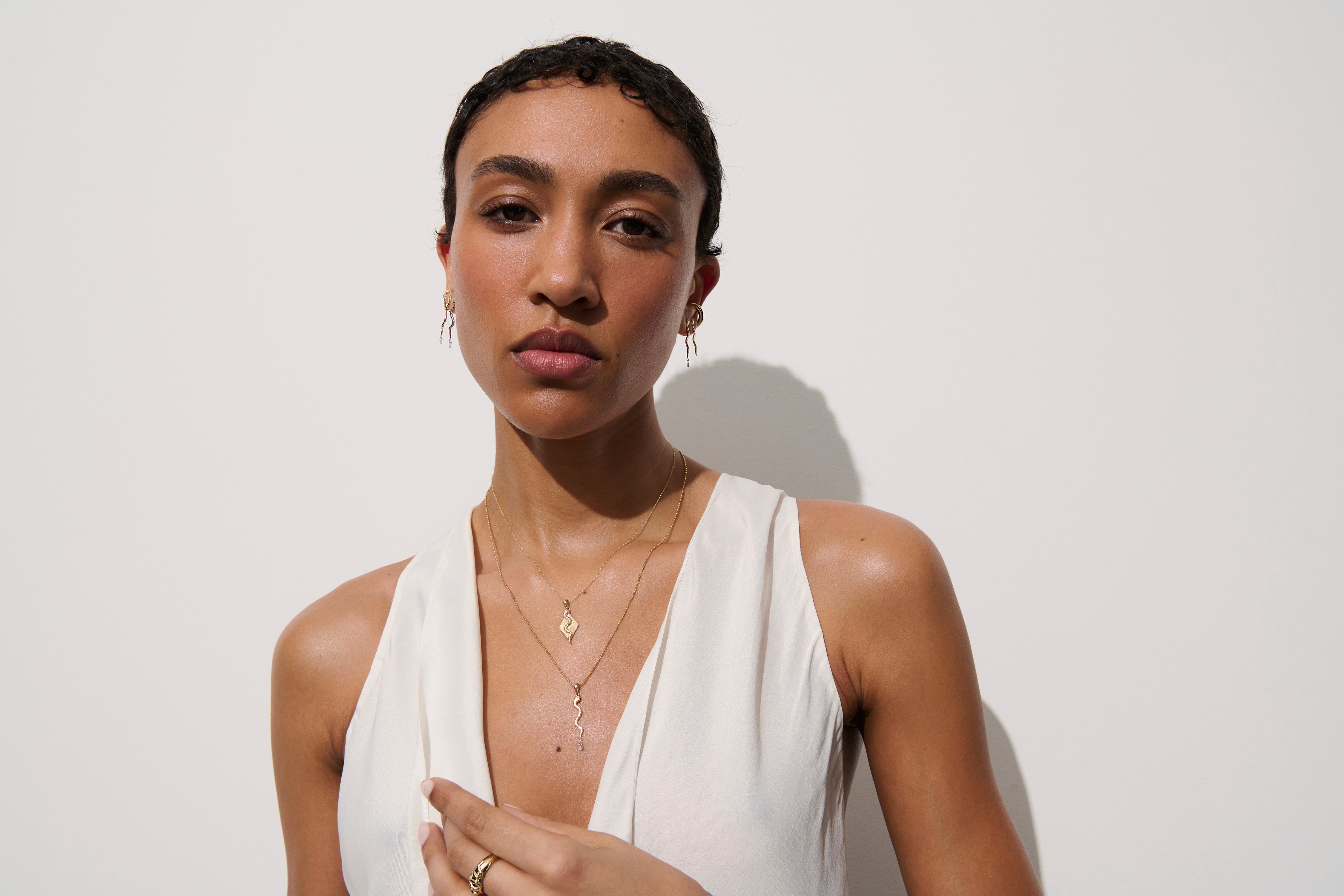 Alt="Vale Jewelry Model wearing textured serpent earrings with diamond drops, textured serpent necklace with diamond drop, textured serpent ray necklace, as well as other eternals collection jewelry"