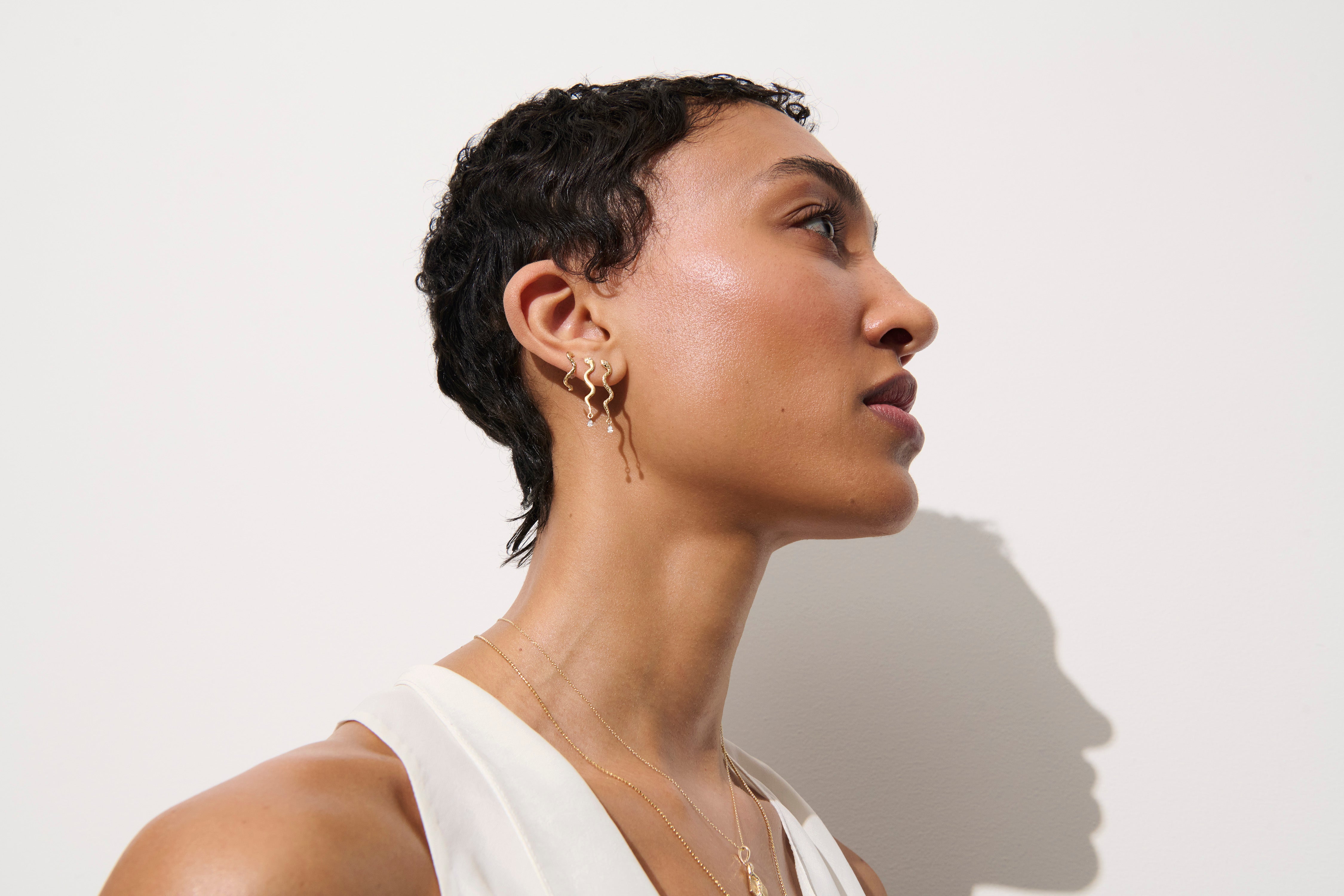 Alt="Vale Jewelry model wearing textured serpent earrings with diamond drops and a few other earrings in profile"
